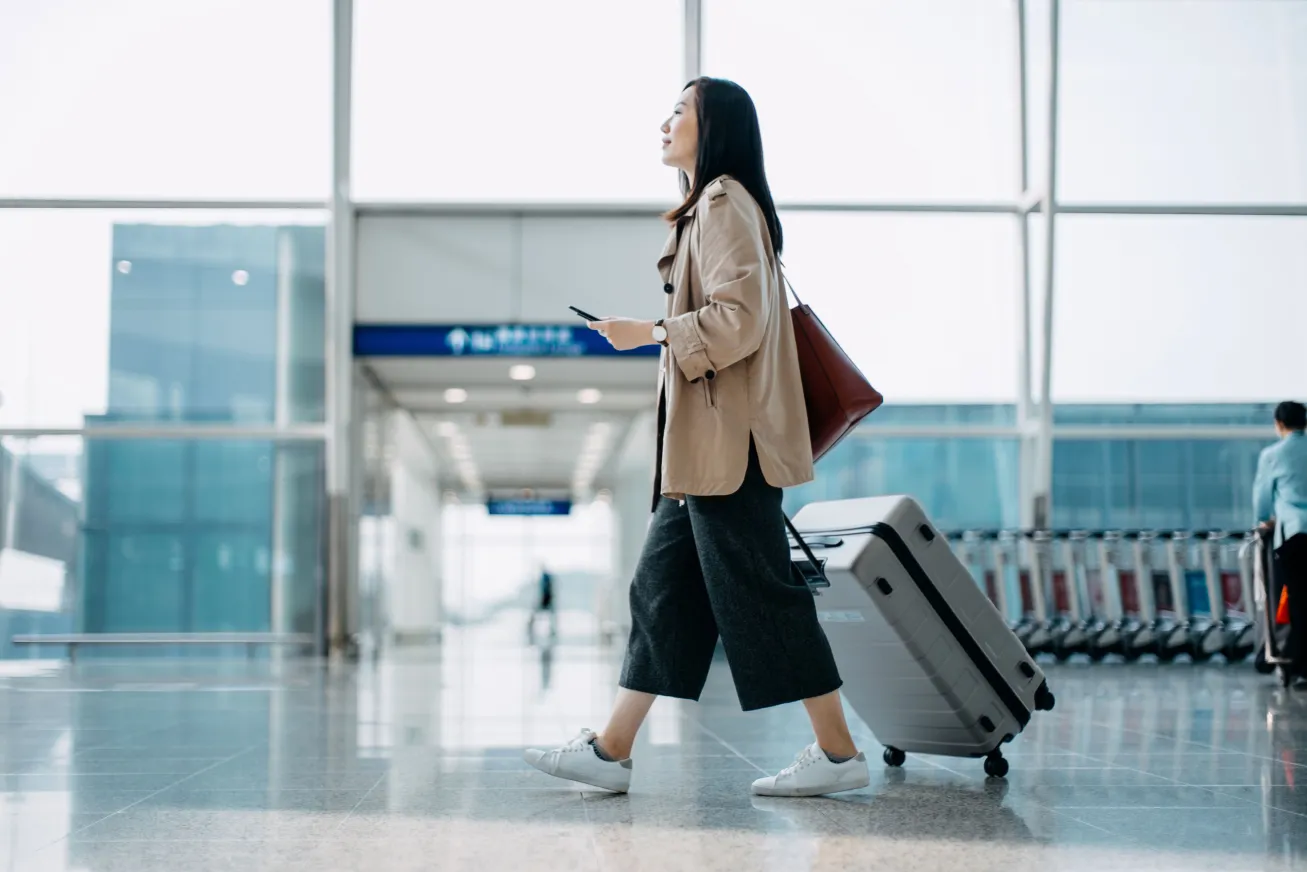Young woman carrying suitcase and holding smartphone on hand, walking in airport terminal.
