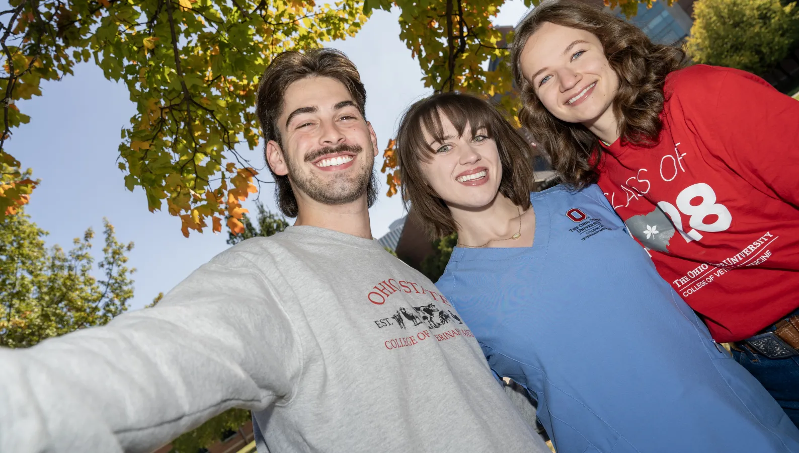 three students, a male in a sweatshirt, female in scrubs and female in class of 2028 tshirt under a tree